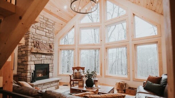 Interior acogedor de una cabaña con chimenea de piedra, ventanas amplias y techo de madera.