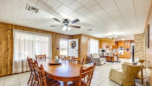 Un grand espace à manger avec une table et des chaises en bois, un ventilateur de plafond et une cuisine en arrière-plan.