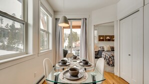 Un coin repas avec une table en verre dressée pour quatre personnes, une vue sur un paysage enneigé par de grandes fenêtres, et un lit avec une couverture à carreaux en arrière-plan.