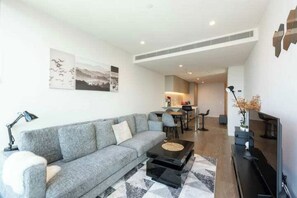 A modern living room with a grey sofa, a coffee table, and a flat-screen TV. There is a dining area in the background with a kitchen visible.