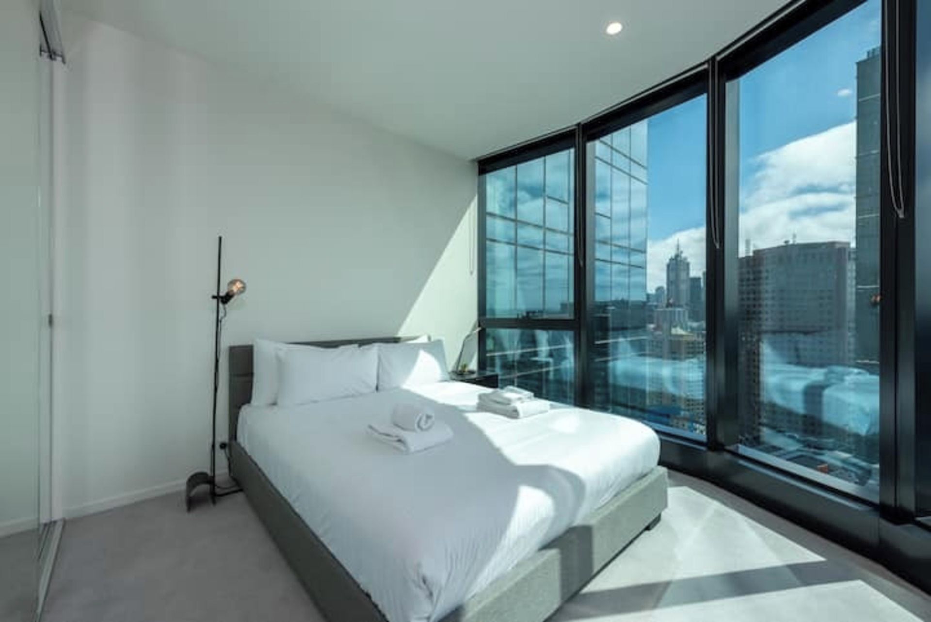 Quarto moderno com uma cama grande, luminária de cabeceira e vista para a cidade através de janelas do chão ao teto.