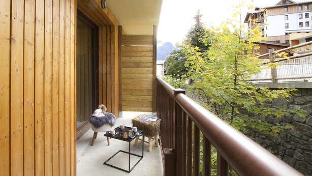 A wooden balcony with a person sitting on a chair, a small table with a cup and a book, and a view of a mountain and buildings in the background. - 300m from ski slopes, 2nd floor, balcony, tv, ski locker, 48m², Les 2 Alpes (Mont-de-Lans)
