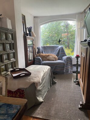 A cozy room with a patterned armchair, a small table with a tray, a bookshelf, and a view of greenery through a large window.