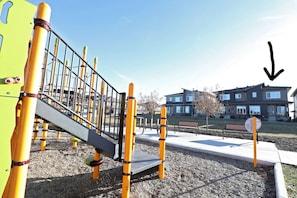 Ein Spielplatz mit Rutsche und Klettergerüsten, umgeben von einer gepflasterten Fläche und einem Gebäude im Hintergrund.