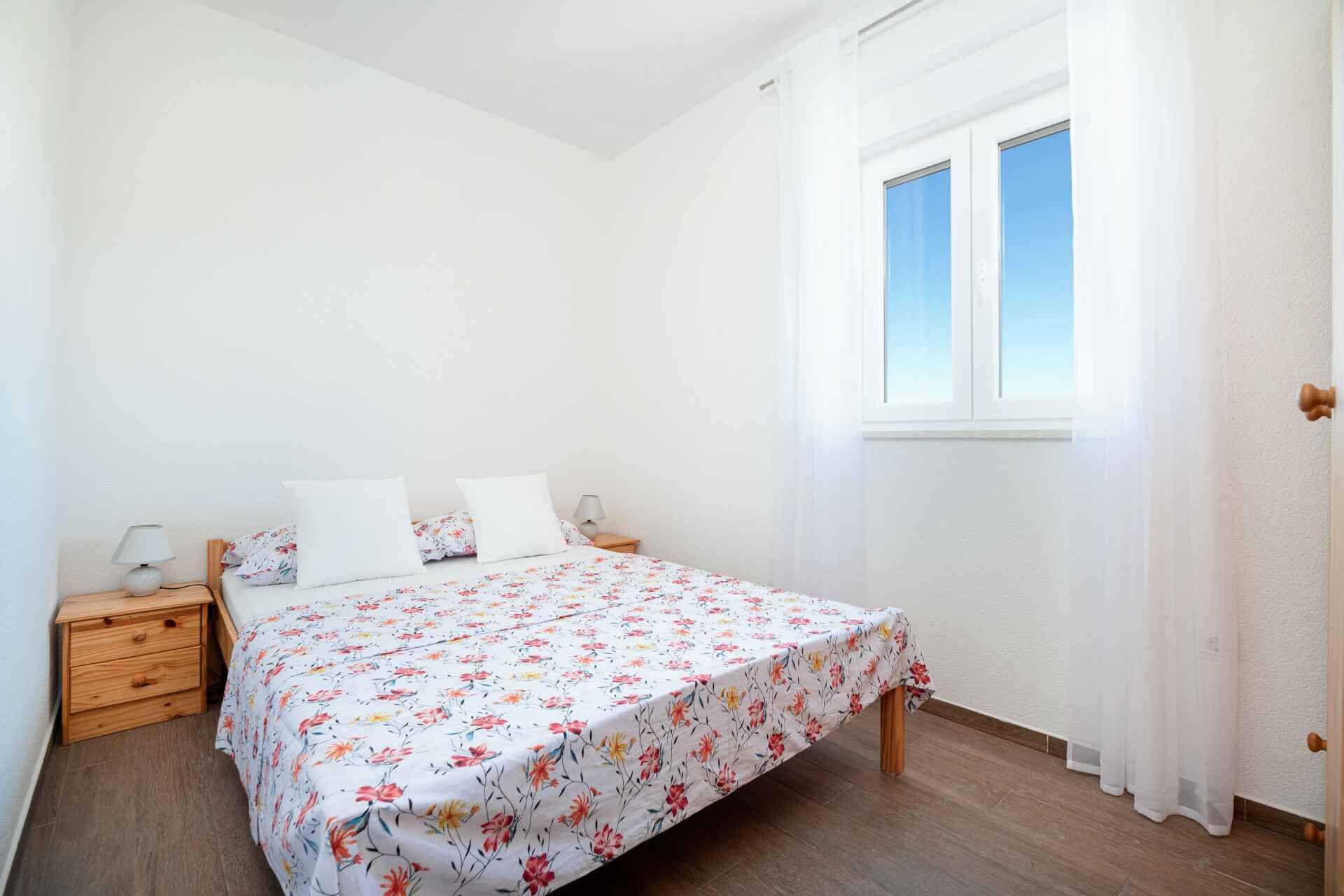 Een slaapkamer met een bed, twee nachtkastjes, een raam met witte gordijnen en een houten vloer.
