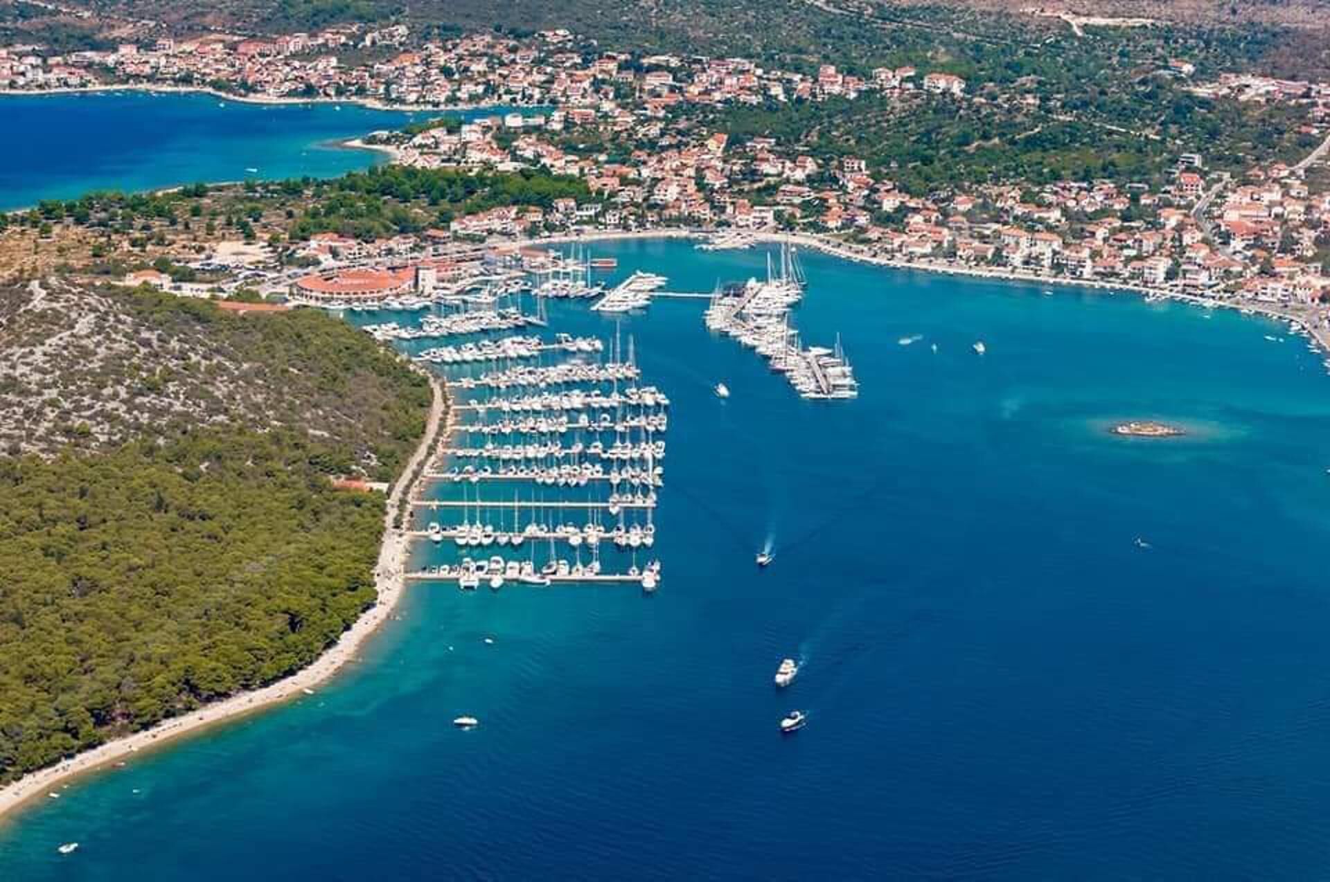 An aerial view of a coastal town with a marina filled with boats, surrounded by residential buildings and a hilly landscape.