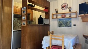 Een compacte keuken met houten kastjes, een ingebouwde magnetron en een kleine koelkast. Er staat een houten tafel met een witte tafelkleed en een bank met een rode kussen. Een televisie is aan de muur boven de tafel bevestigd.