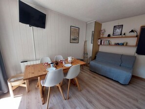 A dining area with a wooden table and white chairs, a grey sofa, and a bookshelf with books and decorative items. - 50m from ski slopes, 3rd floor, balcony, tv, ski locker, 29m², Les 2 Alpes (Les Deux Alpes)