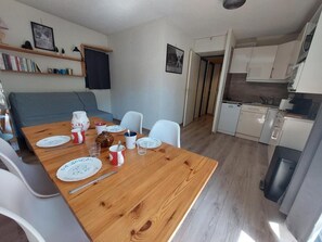 A dining area with a wooden table set for four, white chairs, and a kitchen in the background. - 50m from ski slopes, 3rd floor, balcony, tv, ski locker, 29m², Les 2 Alpes (Les Deux Alpes)
