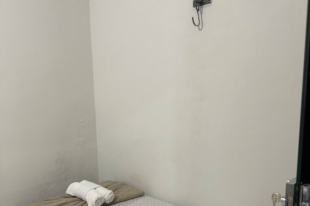 A small, single-bed room with a plain white wall, a bed with a grey checkered bedspread and white pillows, a brown blanket, and a white towel.