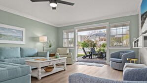 A spacious living room with a light blue sofa, a white coffee table, and a gray armchair. There is a large window with blinds, a ceiling fan, and a view of the outdoors.
