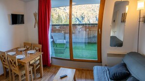A cozy room with a dining table, chairs, a sofa, a TV, and a sliding glass door leading to a balcony with a view of trees. - 200m from ski slopes, 1st floor, view valley/exceptional, swimming pool, balcony, tv, ski locker (Les Orres)