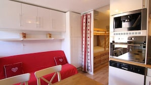 A compact kitchen with white cabinets, a red sofa, a dining table, and a built-in oven.