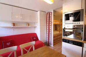 A compact kitchen with white cabinets, a red sofa, a dining table, and a built-in oven.
