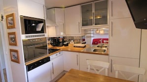 A compact kitchen with white cabinets, a built-in oven, a dishwasher, and a microwave. There is a wooden countertop with a sink and a red chair.