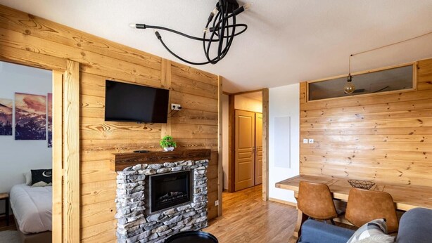 A cozy living room with a stone fireplace, wooden walls, a flat-screen TV, and a dining area with chairs. - 50m from ski slopes, 2nd floor, fitness, swimming pool, sauna, balcony, fireplace or stove, tv (Les Orres)
