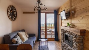 A cozy living room with a stone fireplace, a grey sofa, and a wooden dining table. - 50m from ski slopes, 2nd floor, fitness, swimming pool, sauna, balcony, fireplace or stove, tv (Les Orres)