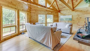 A wooden cabin with a large bed, a sofa, and a dining table. There are large windows and a view of the outdoors. - Pocono Dog-Friendly Home on River (Damascus)