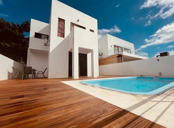 A modern house with a swimming pool, wooden deck, and a dining area with chairs.