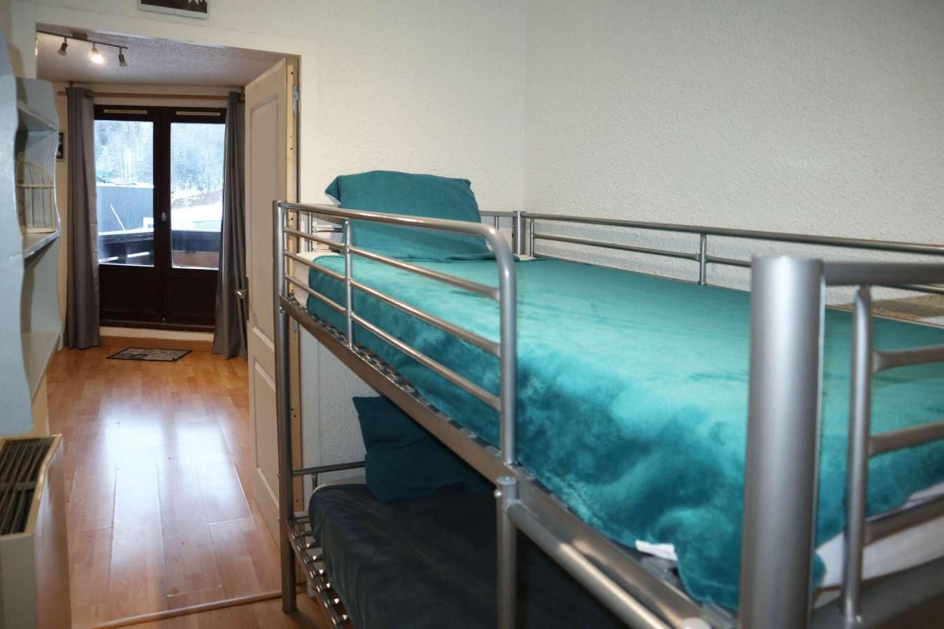 Un lit superposé avec un matelas de couleur turquoise dans une pièce dont le sol est en bois et qui donne sur une zone extérieure par une porte.