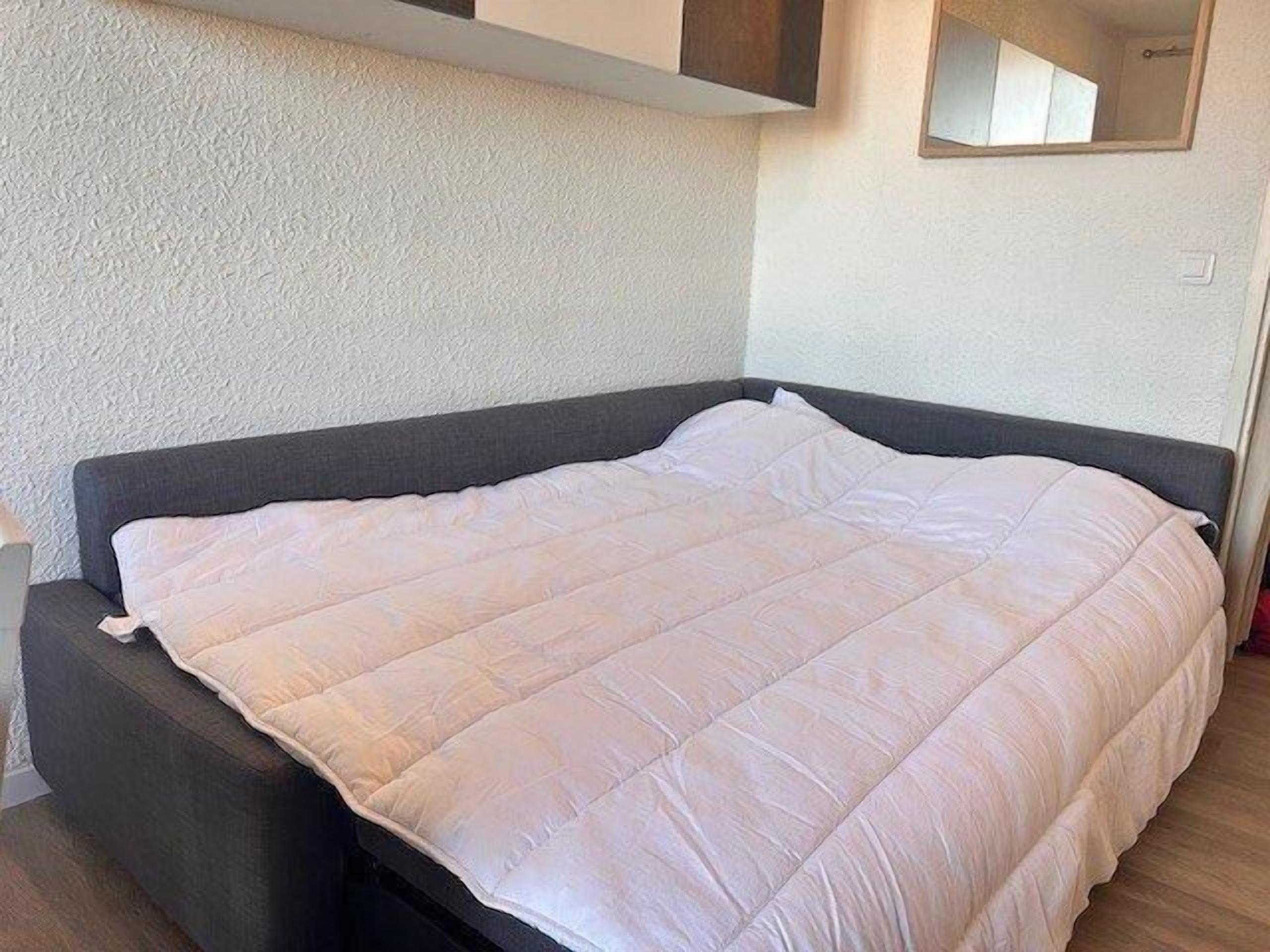 A bed with a white quilt in a room with a textured wall and a small mirror above it.