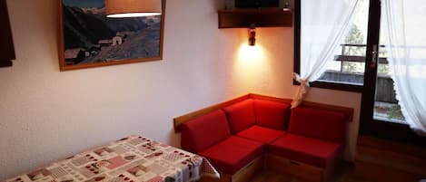 A cozy room with a red sofa, a table with a patterned tablecloth, a hanging lamp, a mounted television, and a window with curtains.
