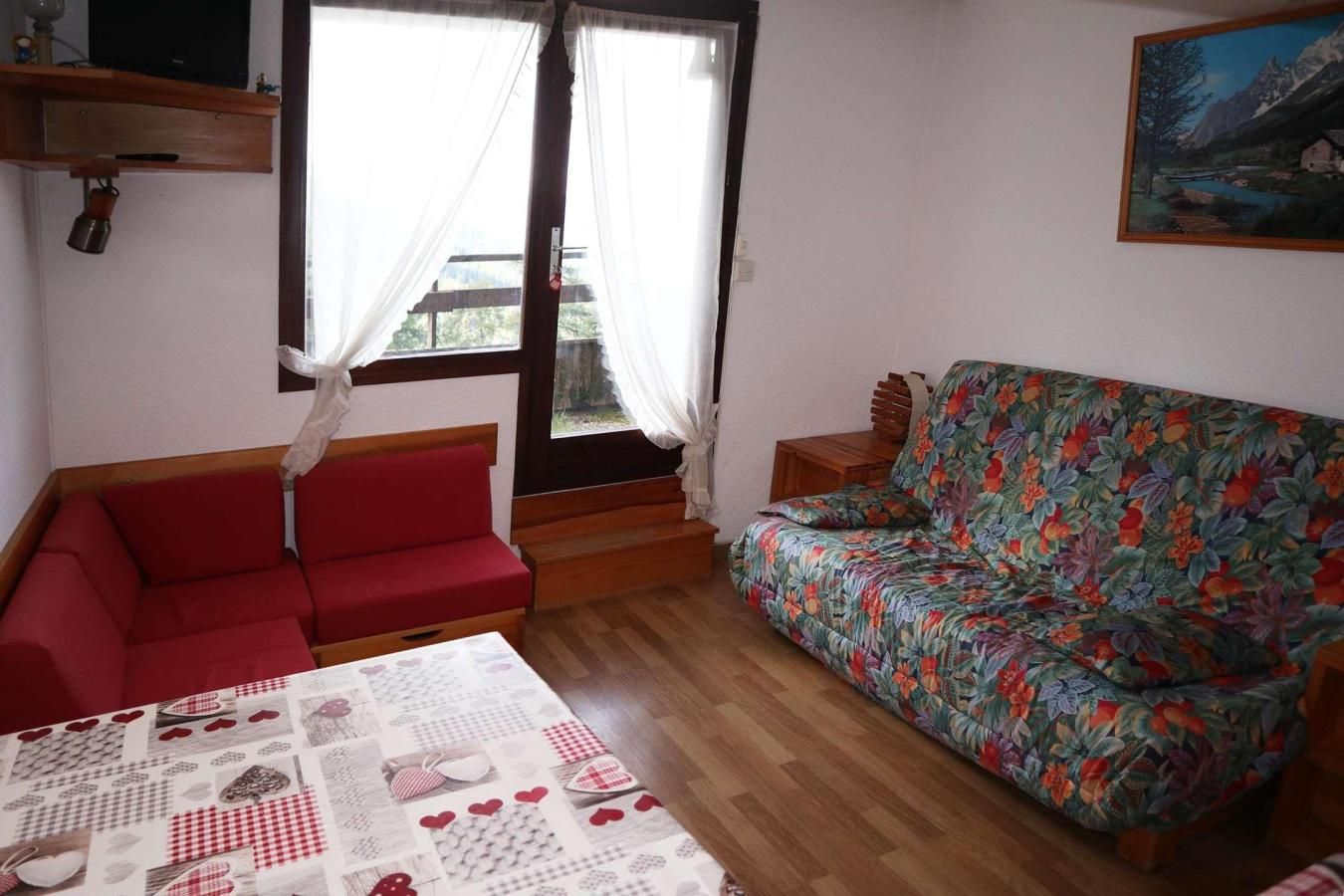 A room with a red sofa, a floral-patterned sofa, a wooden table with a patterned tablecloth, and a painting on the wall.