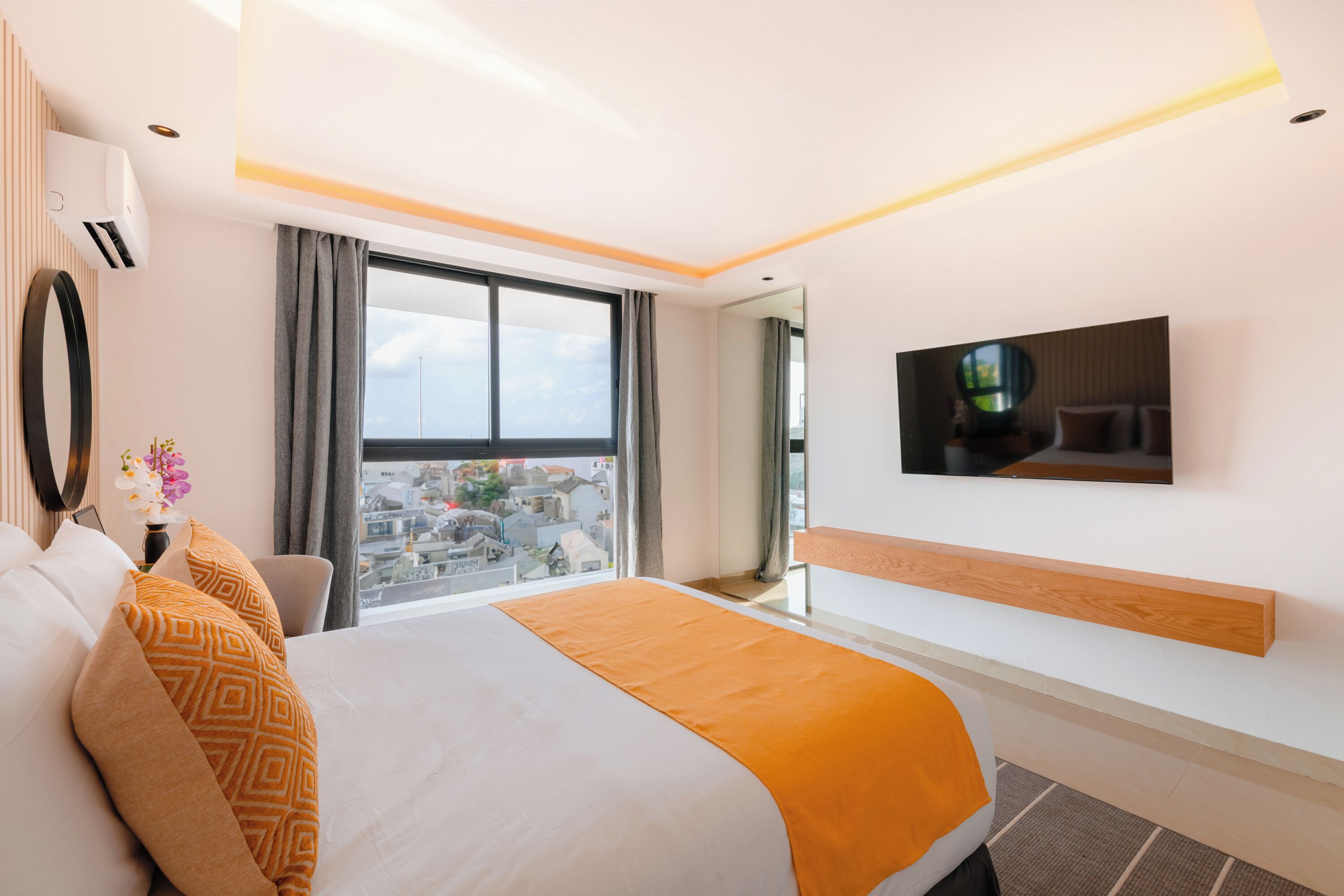 A modern hotel room with a large bed, a TV mounted on the wall, and a view of the city through the window.
