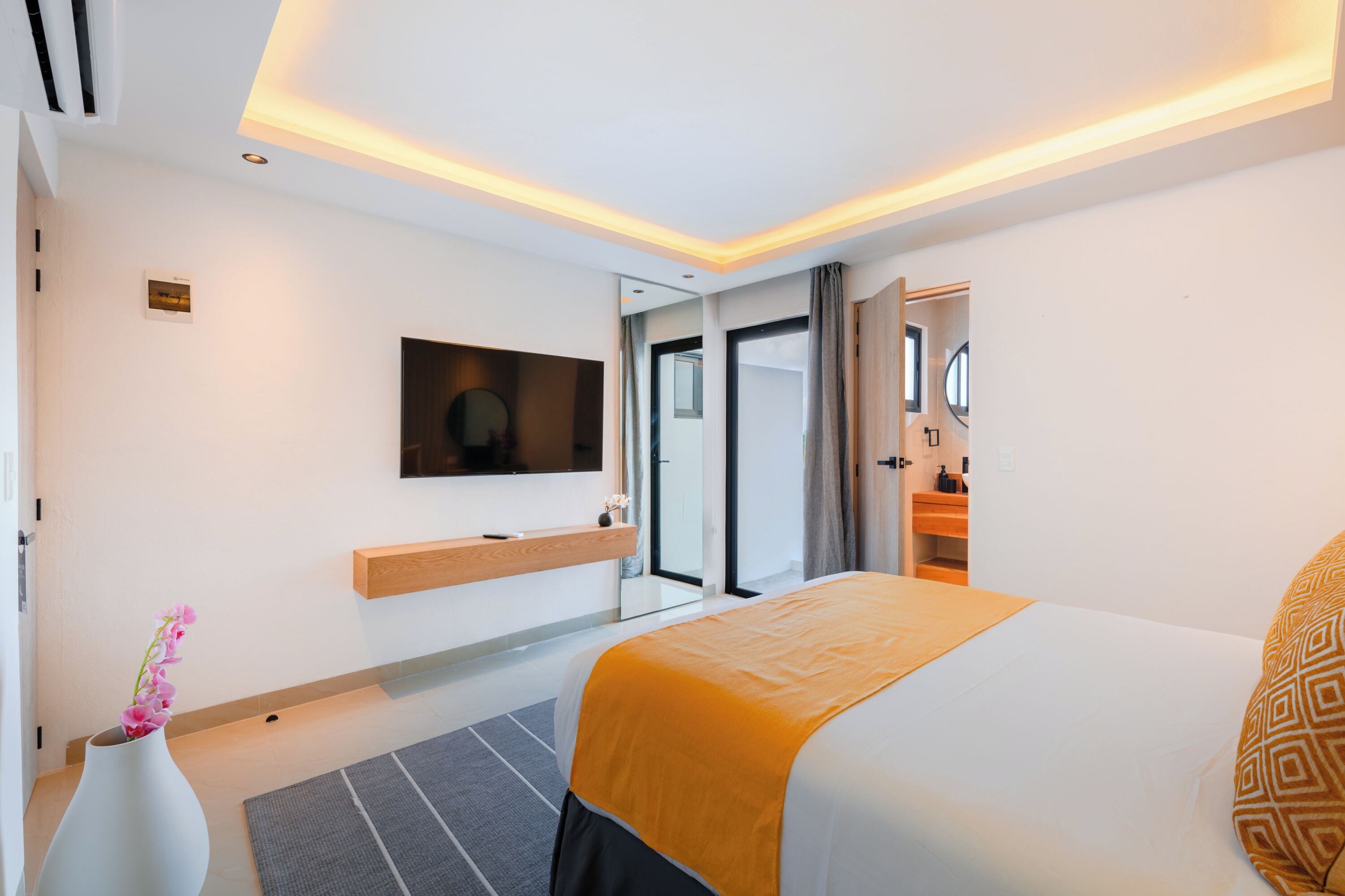 Una habitación de hotel moderna con una cama grande, un televisor de pantalla plana en la pared, una mesita de noche y un pequeño jarrón con flores.