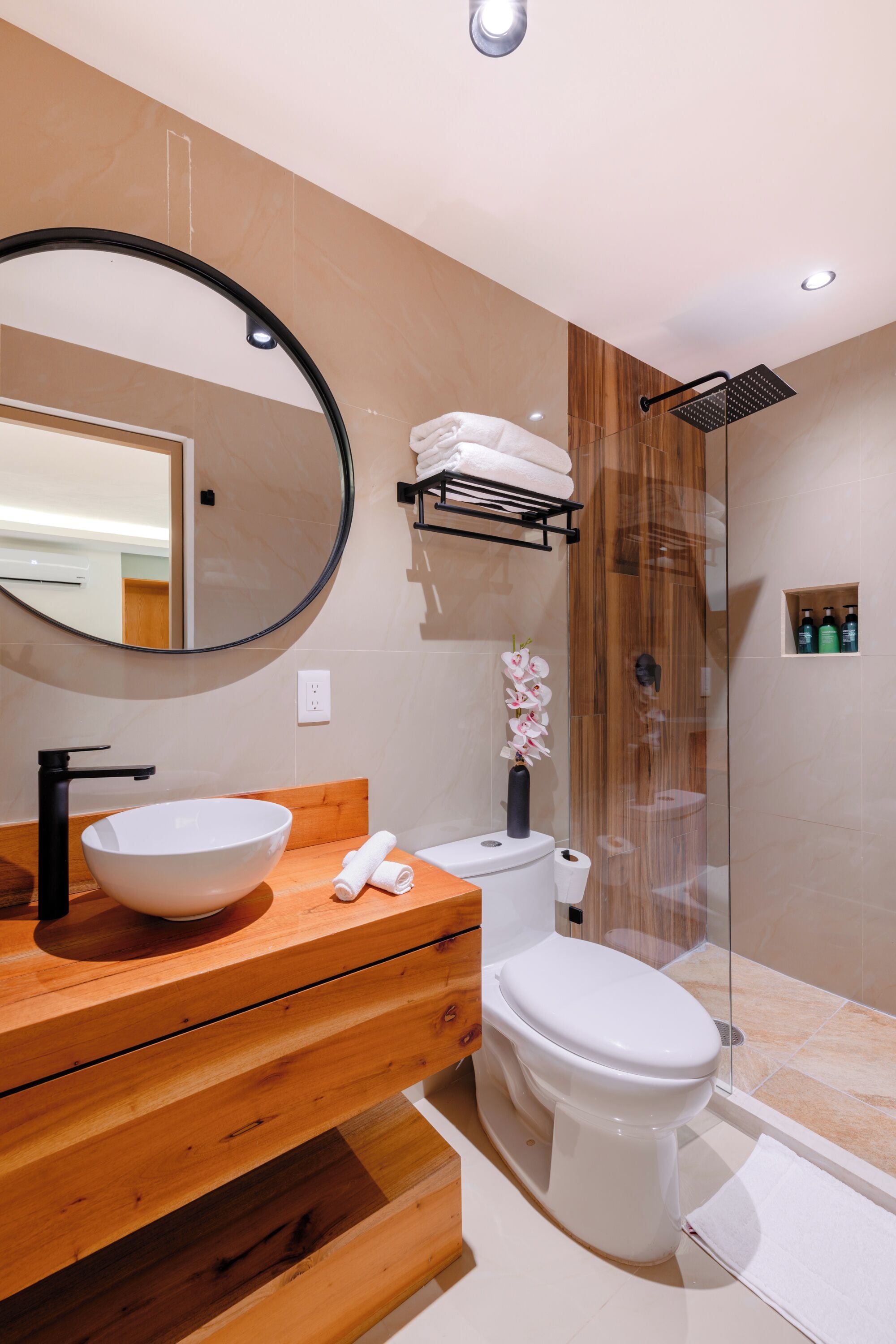 Un baño moderno con tocador de madera, un espejo redondo, un lavabo blanco, un inodoro y una zona de ducha con mampara de cristal.
