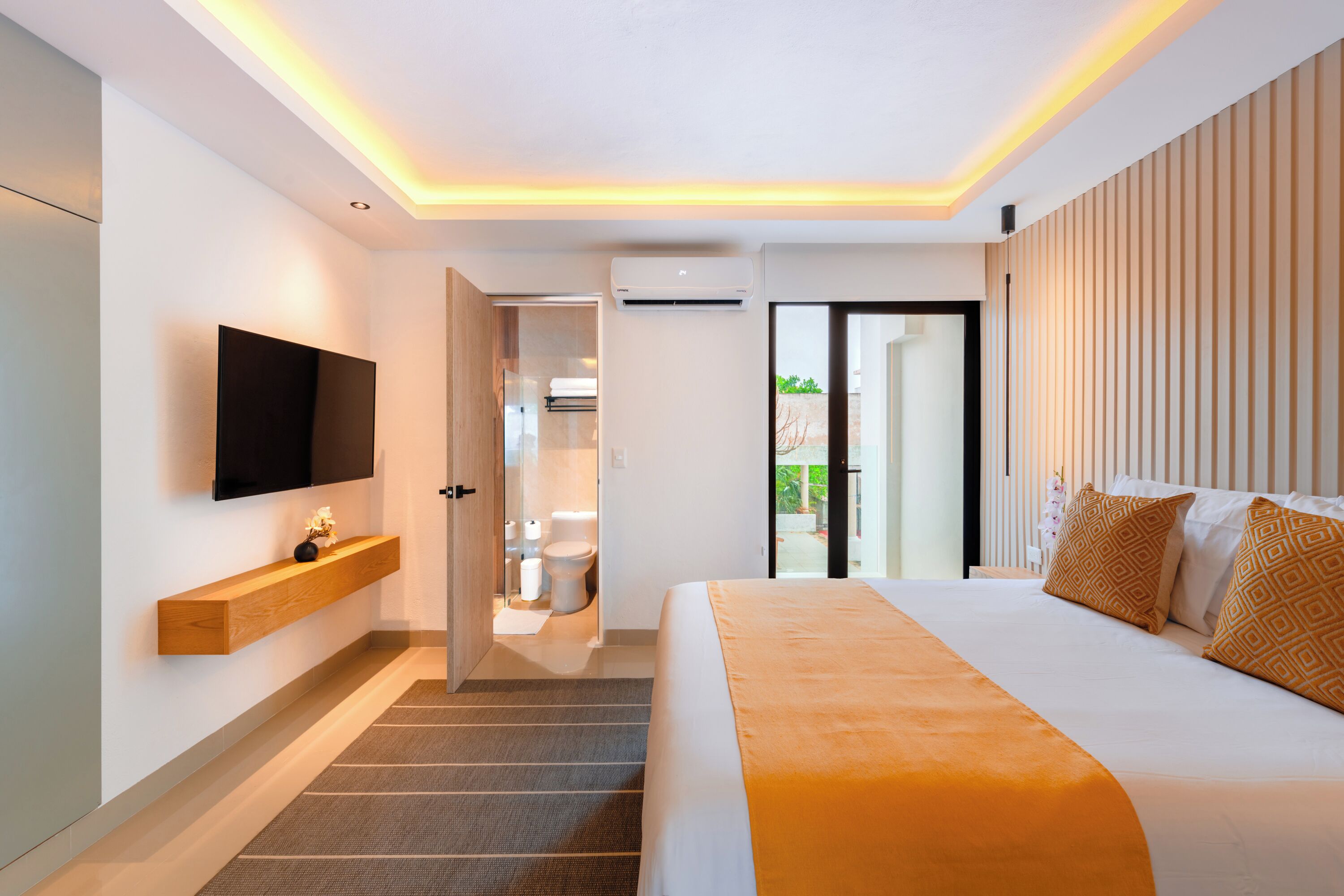 A modern hotel room with a large bed, a TV mounted on the wall, a wooden shelf, and a bathroom visible through an open door.