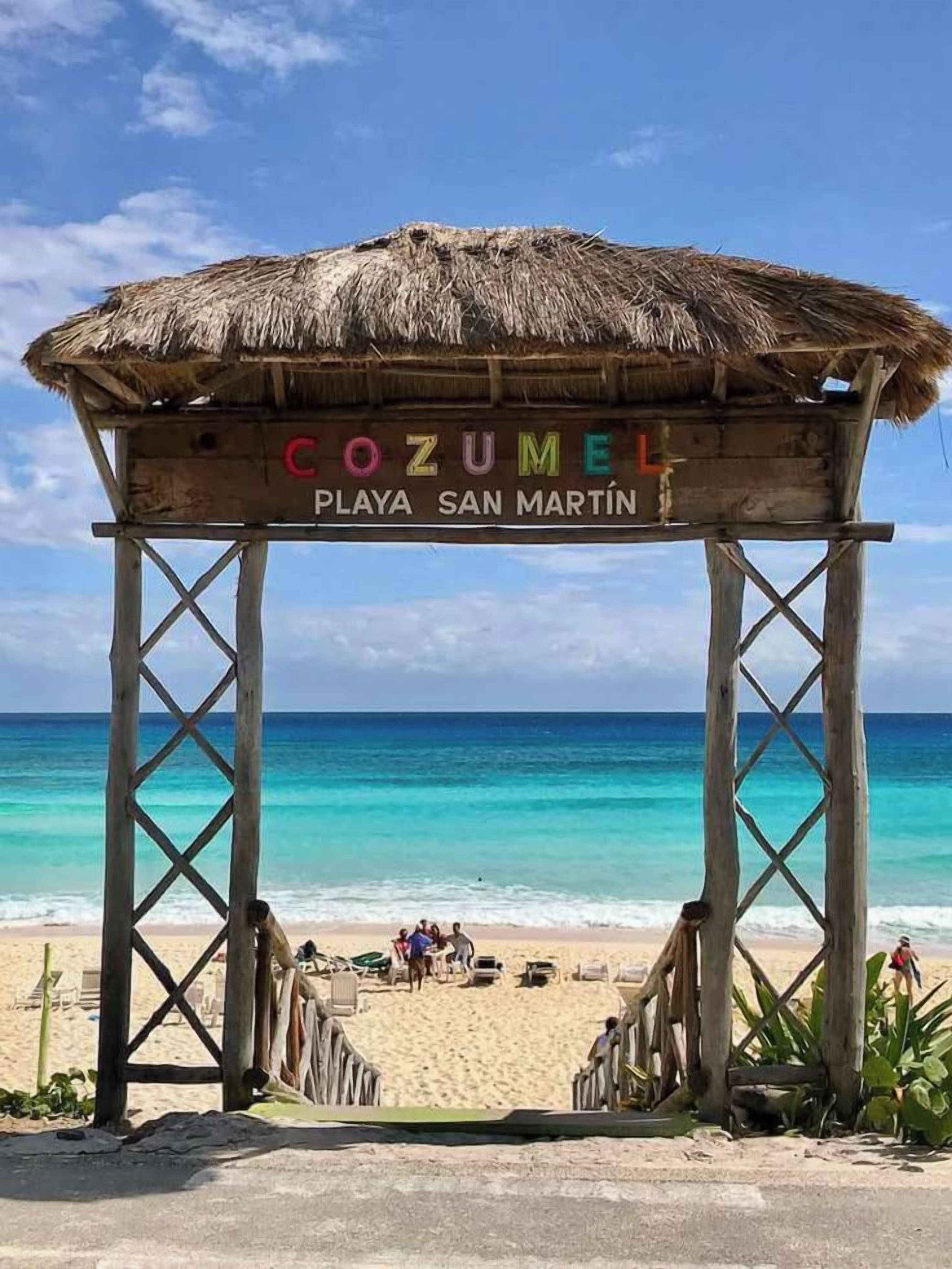 Una estructura de madera con techo de paja identificada con 'COZUMEL PLAYA SAN MARTIN' en una playa.