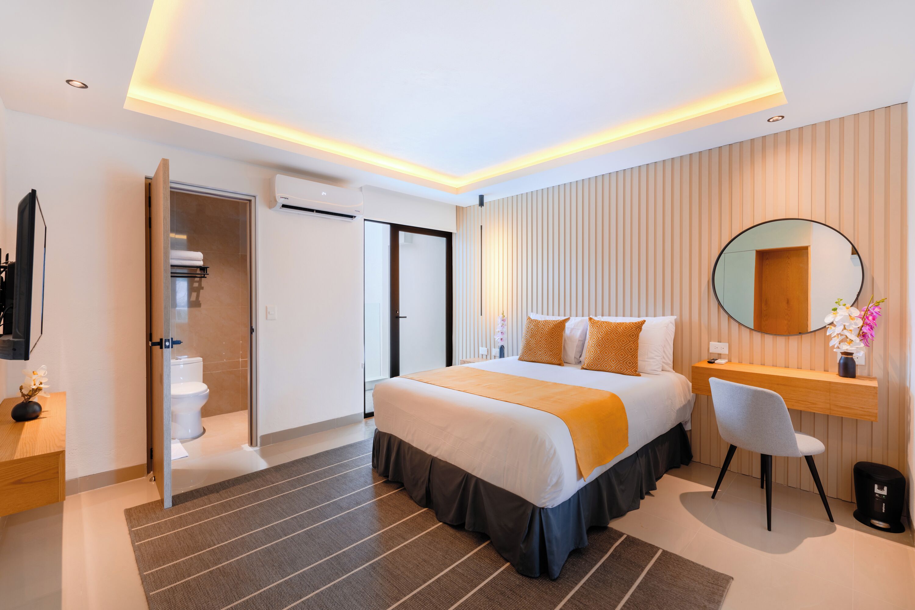 A modern hotel room with a large bed, a desk, a chair, a mirror, and a bathroom visible through an open door.