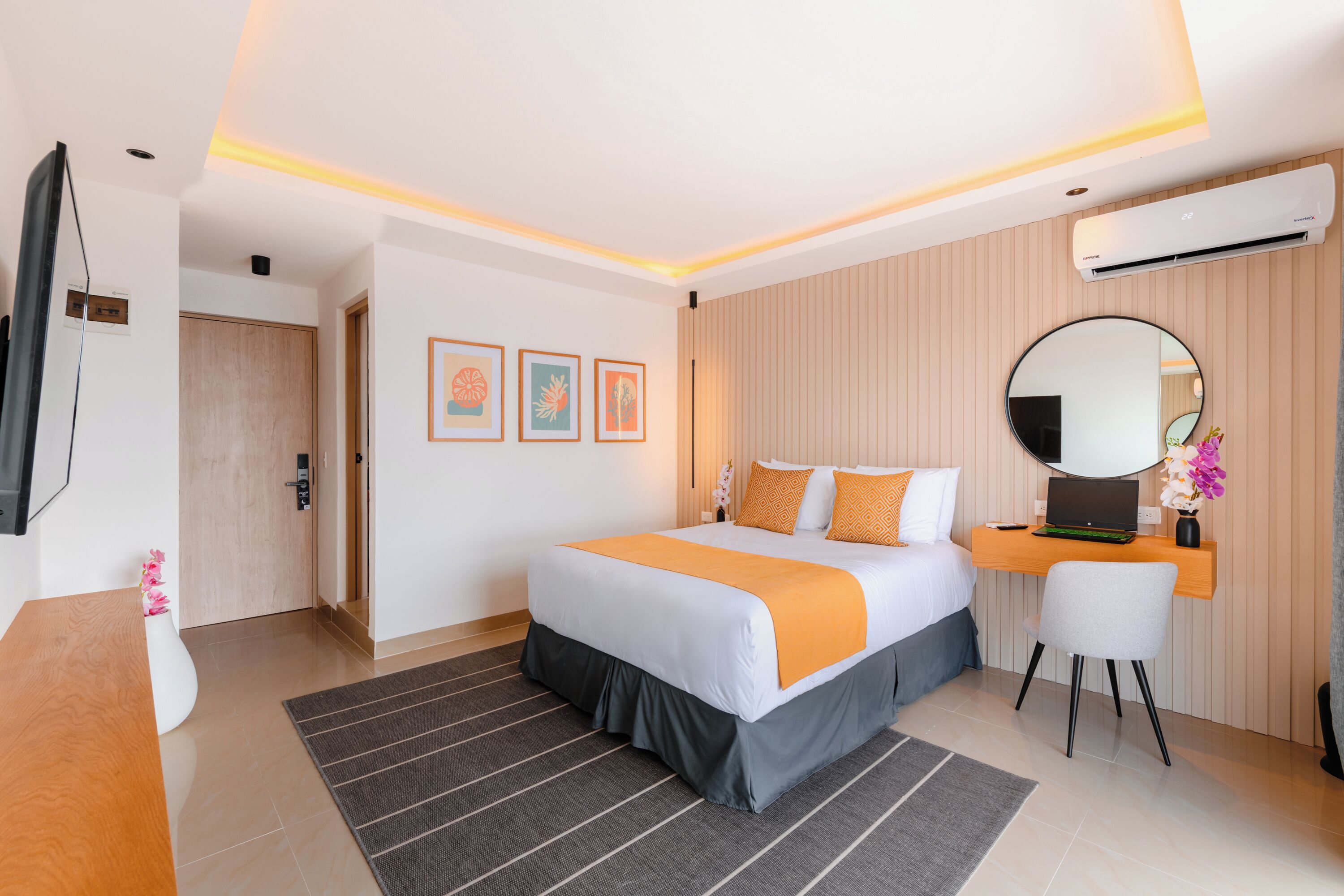 A modern hotel room with a large bed, a desk with a laptop, a round mirror, and a flat-screen TV.