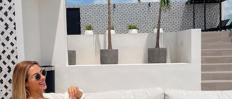 A woman relaxing on a white outdoor sofa with a drink, surrounded by palm trees and a patterned wall.