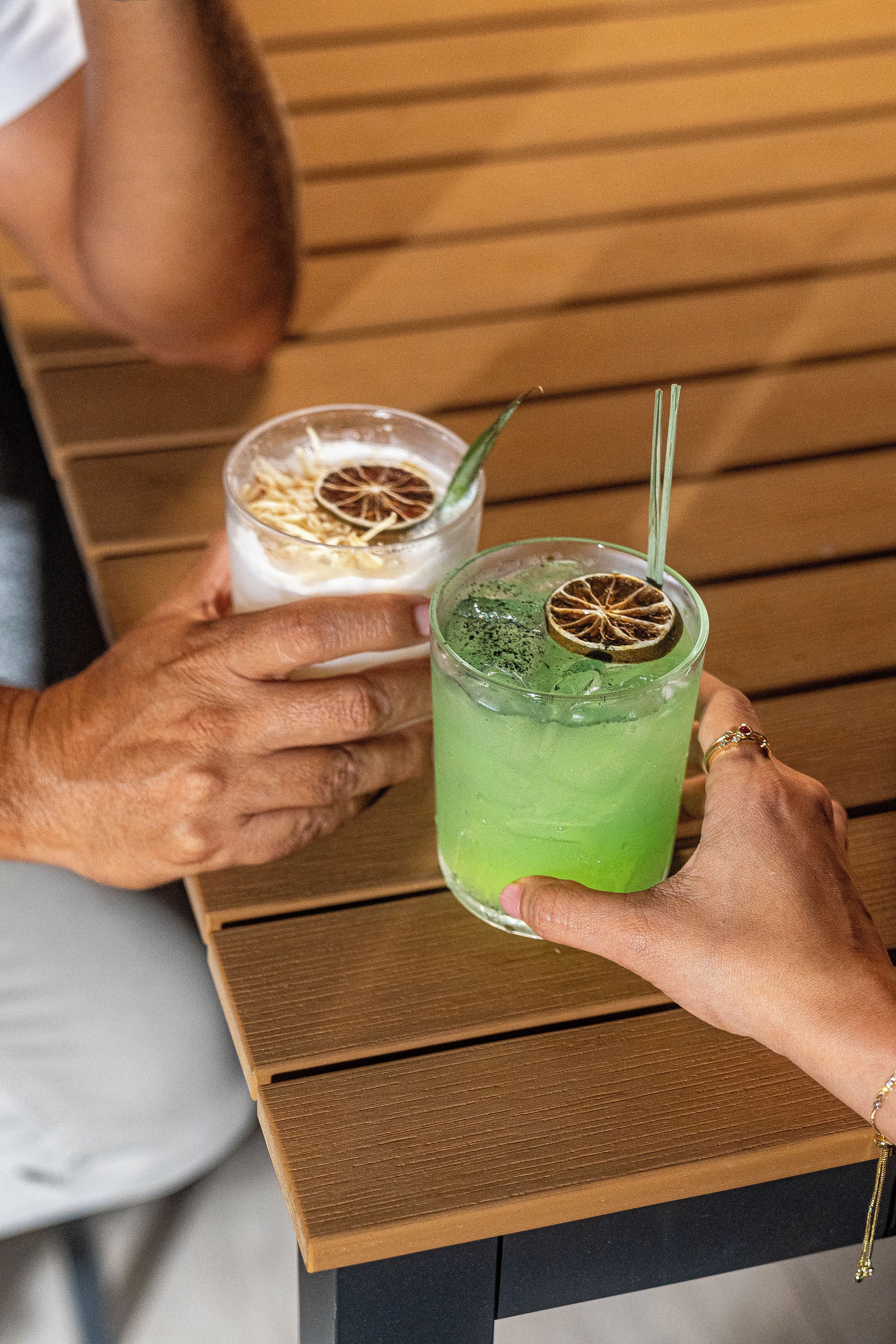 Deux personnes tiennent des verres contenant des boissons, chacun muni d’une paille : l’un contient une boisson verte et l’autre une boisson claire avec une tranche d’orange.