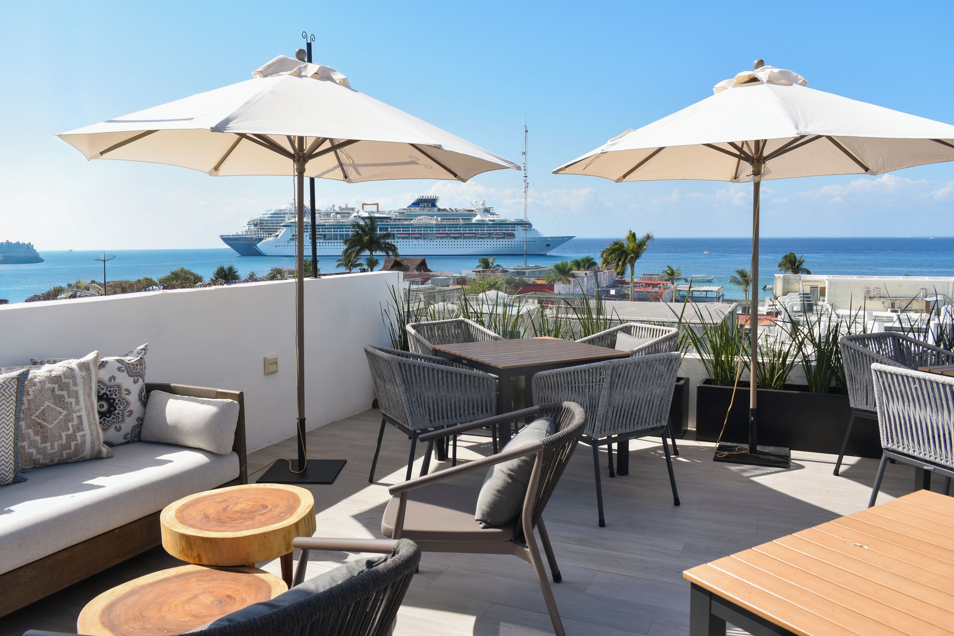 A terrace with outdoor seating, a view of the sea, and a cruise ship in the background.