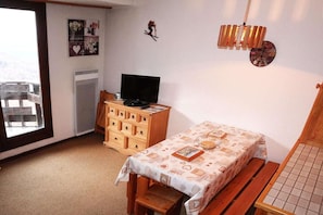 A room with a wooden dresser, a television on top, a small table with a patterned tablecloth, a window with a view, and a wall clock.