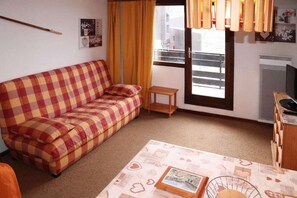 A small, cozy room with a checkered sofa, a small table with a picture frame, a window with curtains, and a radiator.