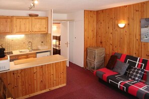 A compact kitchen with wooden cabinets, a tiled backsplash, and a small island. There is a red sofa with patterned cushions and a wall-mounted light fixture. - Resort center, 6th floor, view ski slopes, balcony, 20m², Les Orres (Les Orres)