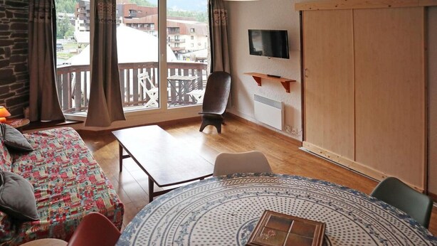 Een kamer met een houten vloer, een ronde tafel met een tafelkleed met dessin, een bank met een kleurrijk kleed, een stoel, een aan de muur bevestigde televisie en een balkon met houten hekwerk.