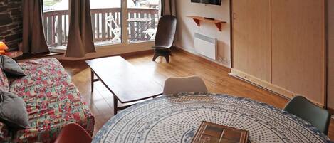 A room with a wooden floor, a round table with a patterned tablecloth, a sofa with a colorful blanket, a chair, a TV mounted on the wall, and a balcony with a wooden railing.