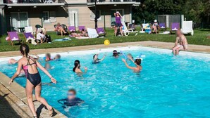 Une piscine avec des personnes dans l’eau et sur le bord, entourée de chaises longues et avec un bâtiment en arrière-plan.
