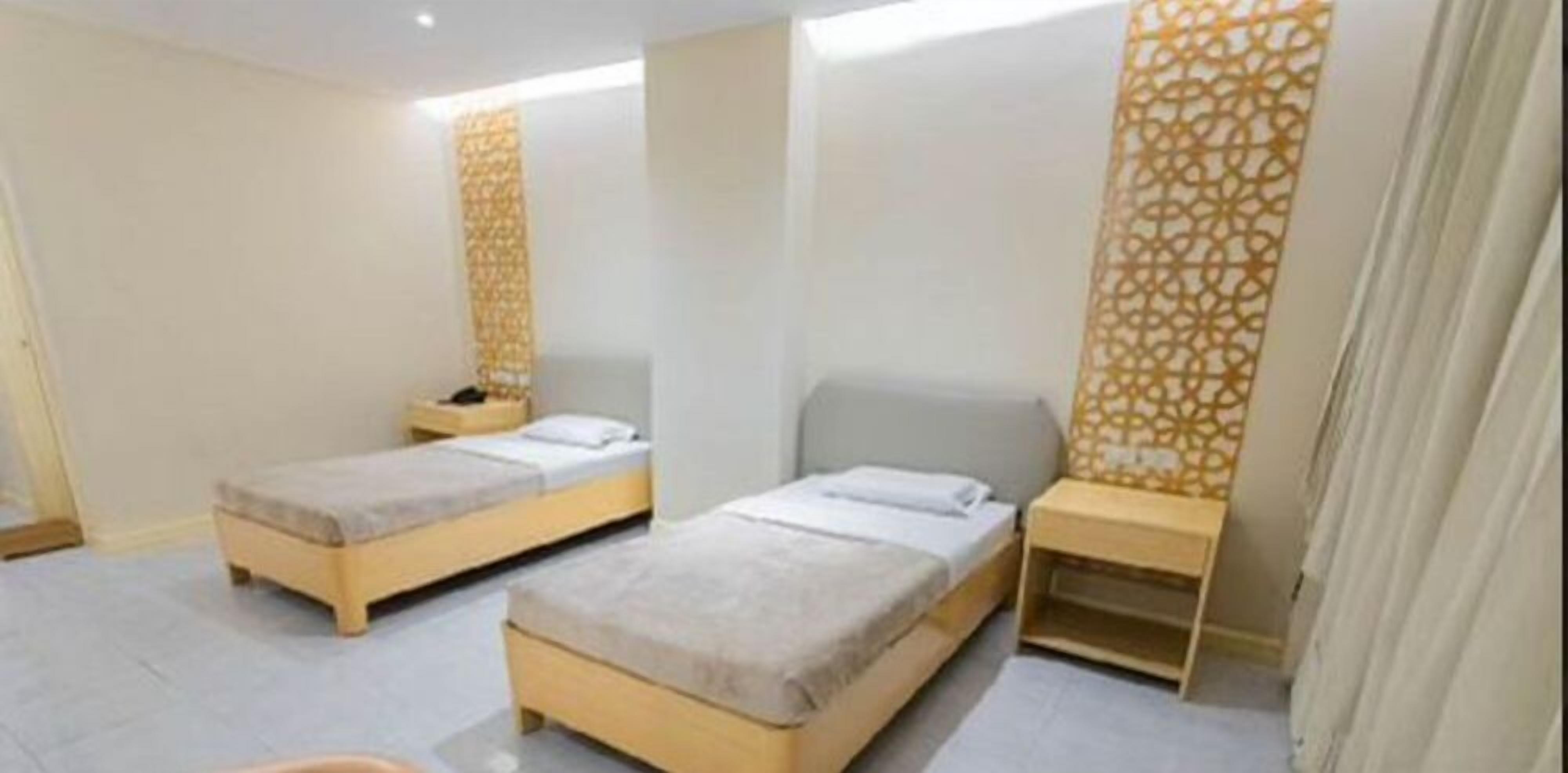 A modern hotel room with two single beds, a small bedside table with a lamp, and a decorative wall panel.