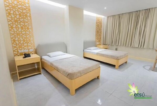 Habitación de hotel con dos camas individuales, una mesita de noche pequeña con lámpara, un panel decorativo en la pared y un ventanal con cortinas.
