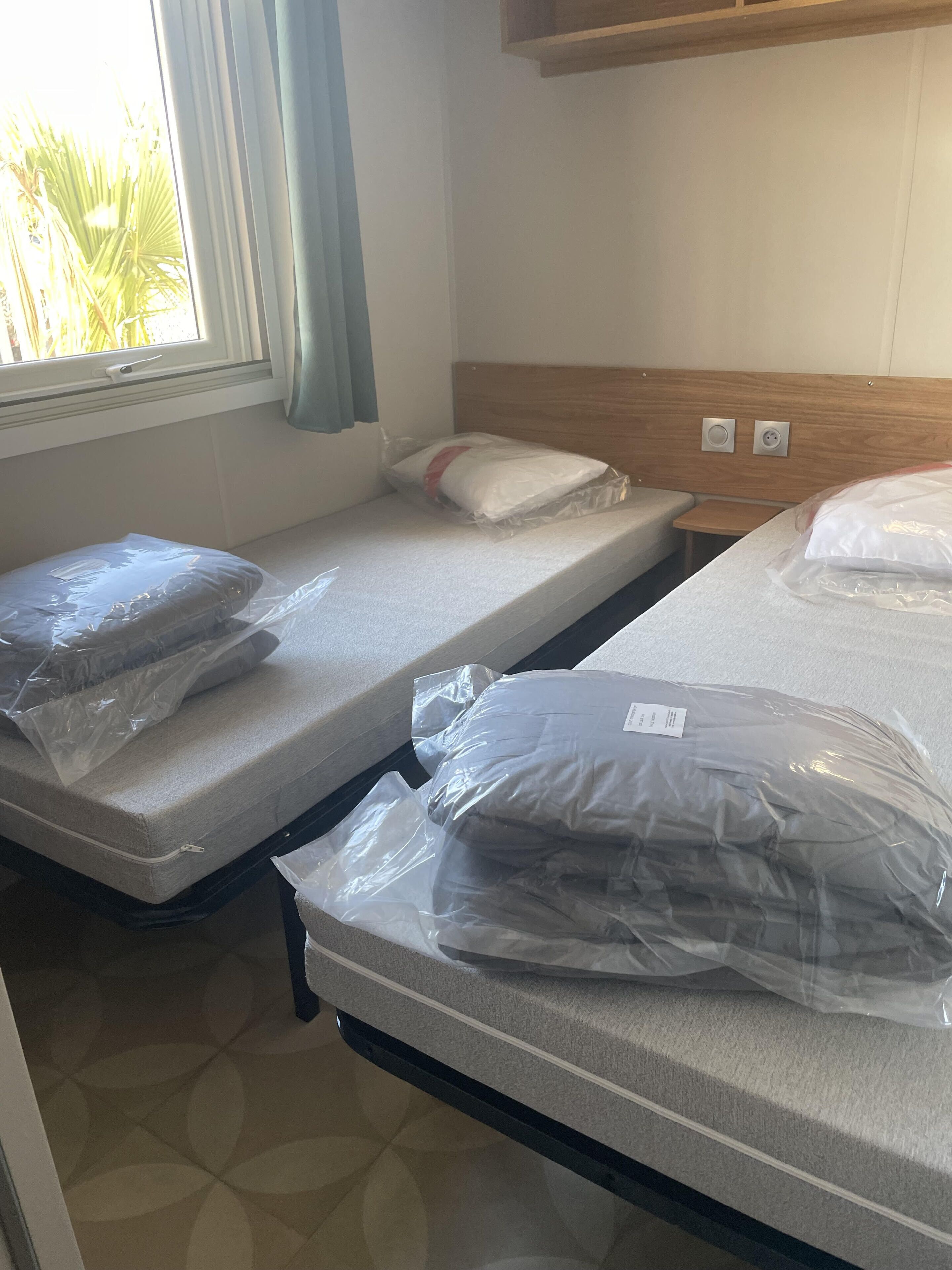Habitación de hotel con dos camas individuales, cada una con un colchón y almohada cubiertos de plástico. Se ve una ventana con cortina, una mesita de noche y un cabecero de madera.