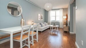 A modern living room with a white sofa, a dining table with chairs, a round mirror, and a large window with curtains. - Du Portage 1-Bedroom Condo (Gatineau)