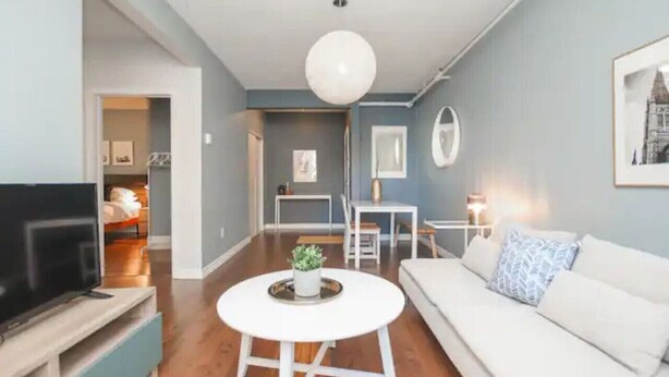A modern living room with a flat-screen TV, a white sofa, a round coffee table, and a dining area with a table and chairs. - Du Portage 1-Bedroom Condo (Gatineau)