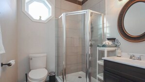 A bathroom with a glass-enclosed shower, a toilet, a sink with a mirror, and a small window. - Du Portage 1-Bedroom Condo (Gatineau)