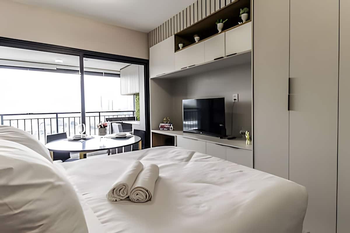 A hotel room with a bed, a TV on a shelf, a dining table with chairs, and a balcony with a railing.